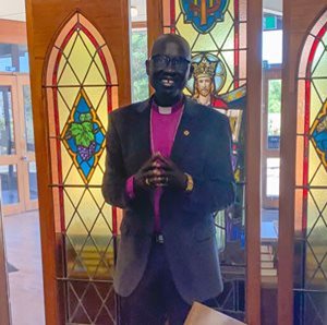 Bishop Jacob Deng Garang Akech standing in front of stained glass windows smiling